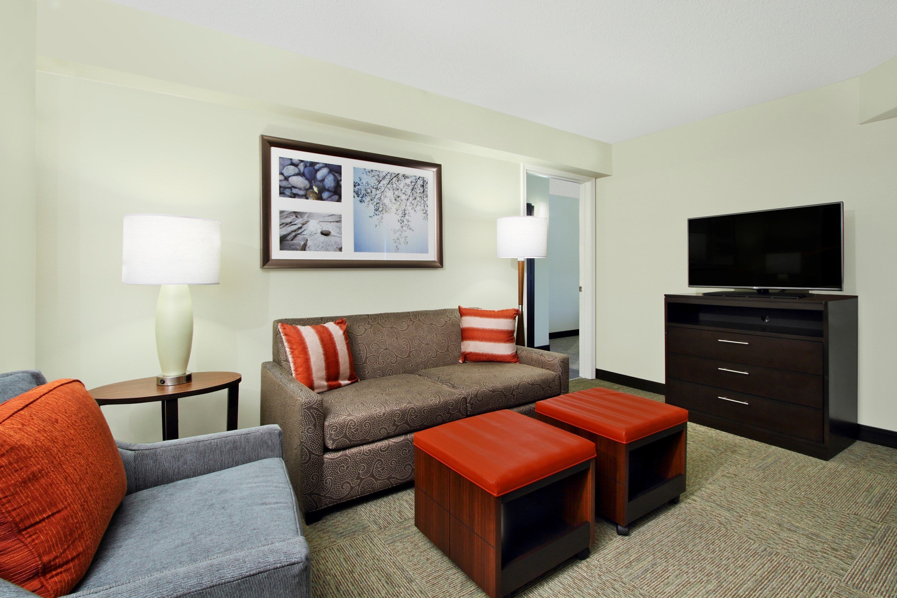 A hotel room with a sofa, two ottomans, a TV, a lamp, and a framed picture on the wall.