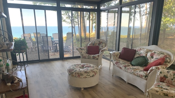 Une pièce baignée de lumière, avec une grande porte vitrée donnant sur un patio. À l'intérieur, on trouve un canapé en osier agrémenté de coussins à motifs floraux, un pouf assorti et une chaise à structure métallique. Une petite table avec une plante et une étagère en métal sur laquelle sont posés divers objets sont également visibles.
