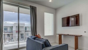A modern living room with a sofa, a TV mounted on the wall, and a balcony with a view of buildings. - Pool View | Bunk Beds | Fast Internet | Loft 32 (Saint George)