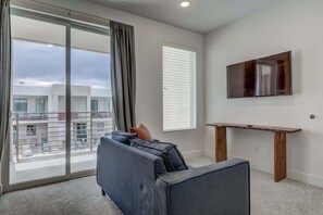 A modern living room with a sofa, a TV mounted on the wall, and a balcony with a view of buildings. - Pool View | Bunk Beds | Fast Internet | Loft 32 (Saint George)