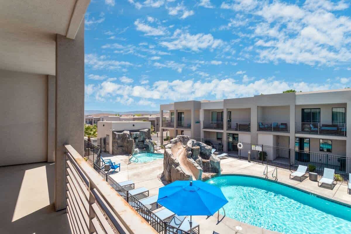 A hotel pool area with a clear sky, a pool, lounge chairs, and a building with balconies.