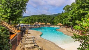 A large outdoor swimming pool with a curved design, surrounded by lounge chairs and umbrellas, set against a backdrop of lush greenery and hills.