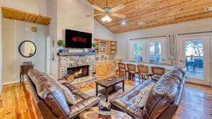 A spacious living room with leather sofas, a stone fireplace, wooden ceiling, and a large TV screen.