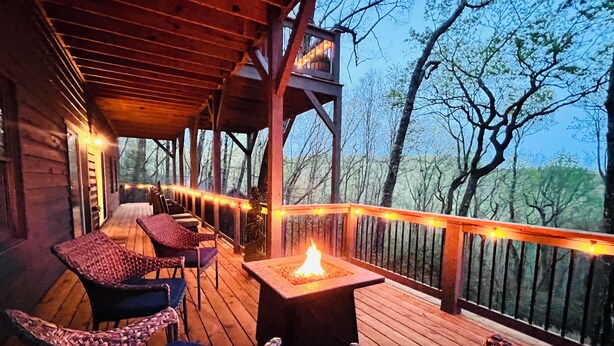 A wooden deck with a fire pit, wicker chairs, and a railing overlooking a forested area.