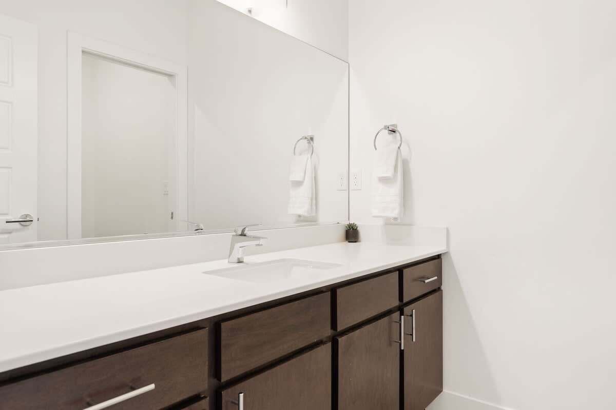 Une salle de bain moderne avec un grand miroir, un lavabo blanc, des meubles en bois foncé et deux serviettes suspendues à des crochets.