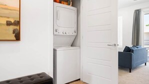 A laundry room with a washing machine and dryer, a bench, and a door leading to a living area with a sofa. - The Lofts #33 | Townhome Pool Resort | Poolside (Saint George)