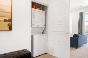 A laundry room with a washing machine and dryer, a bench, and a door leading to a living area with a sofa.