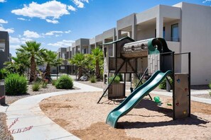 A playground with a slide and climbing wall, surrounded by a sandy area and palm trees, with modern buildings in the background. - The Lofts #33 | Townhome Pool Resort | Poolside (Saint George)