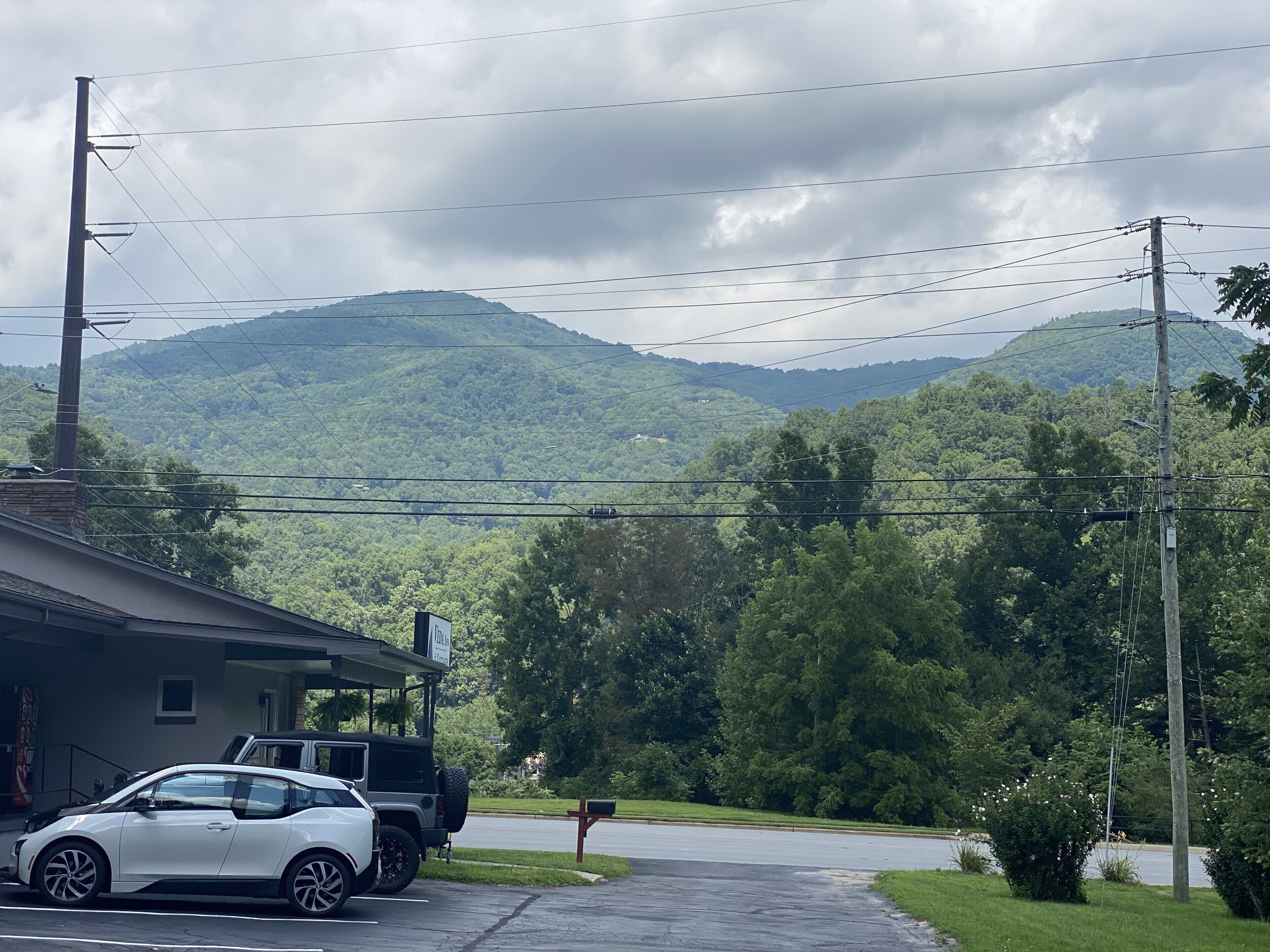 A parking area with a white car and a black SUV. There is a building with a sign on the left. The background features a lush green landscape with hills and a cloudy sky.
