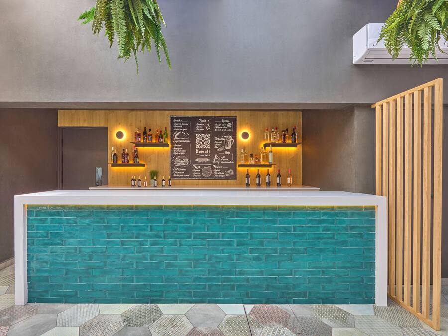 Uma área de bar com balcão de tijolos verde-água, prateleiras com garrafas e um menu em lousa.