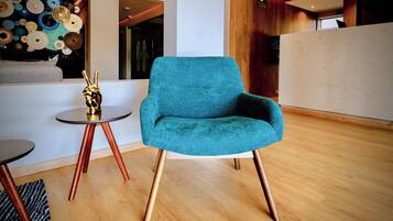 Un sillón azul verdoso con patas doradas, una mesita redonda con una estatua dorada, y un papel tapiz estampado en un espacio interior moderno.