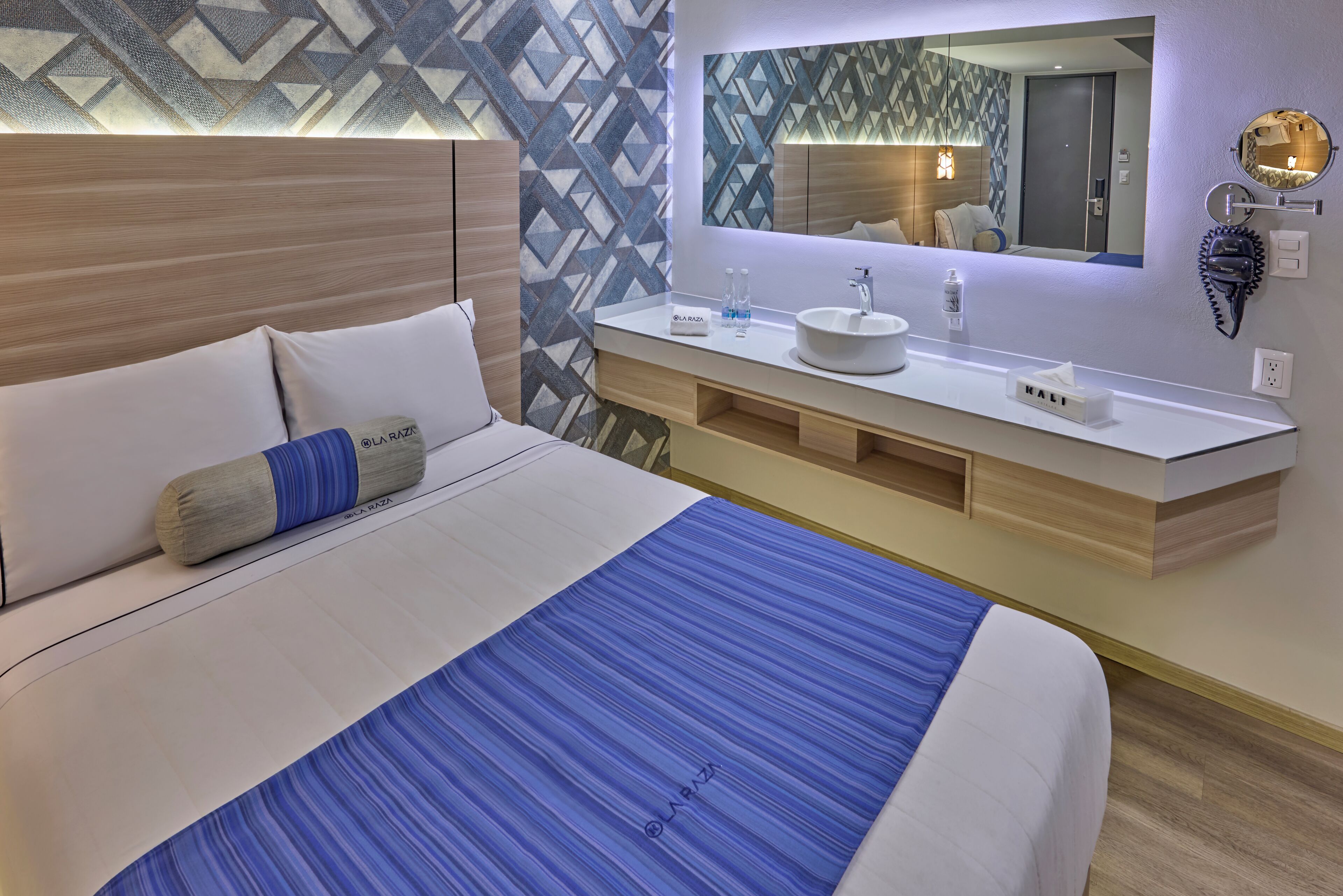 Una habitación de hotel moderna con una cama grande, un cabecero de madera, una colcha a rayas blancas y azules, un tocador blanco con un espejo redondo y una pared con estampado geométrico.