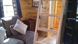 Interior de una cabaña de madera con un sofá, cojines, una escalera, un refrigerador y un cuadro en la pared.