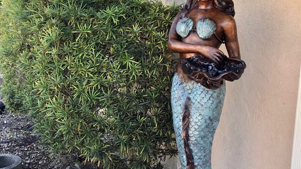Mermaid-patsas, jossa on simpukka, on sijoitettu ulos rakennuksen lähelle. Rakennuksessa on katettu kuisti ja sen edessä kasvaa vihreä pensasaita.