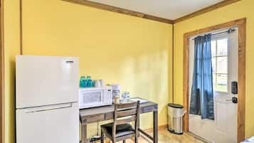 A compact room with a kitchenette, including a refrigerator, microwave, and coffee maker. A chair is placed at a small table, and a door with a curtain is visible.