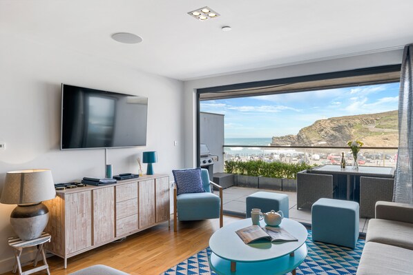 A modern living room with a large TV, comfortable seating, a dining area, and a balcony with a scenic view.