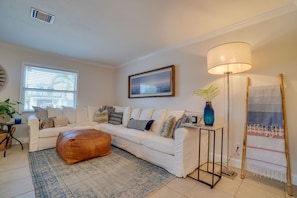 A living room with a white sofa, a brown ottoman, a floor lamp, a painting, and a woven rug.