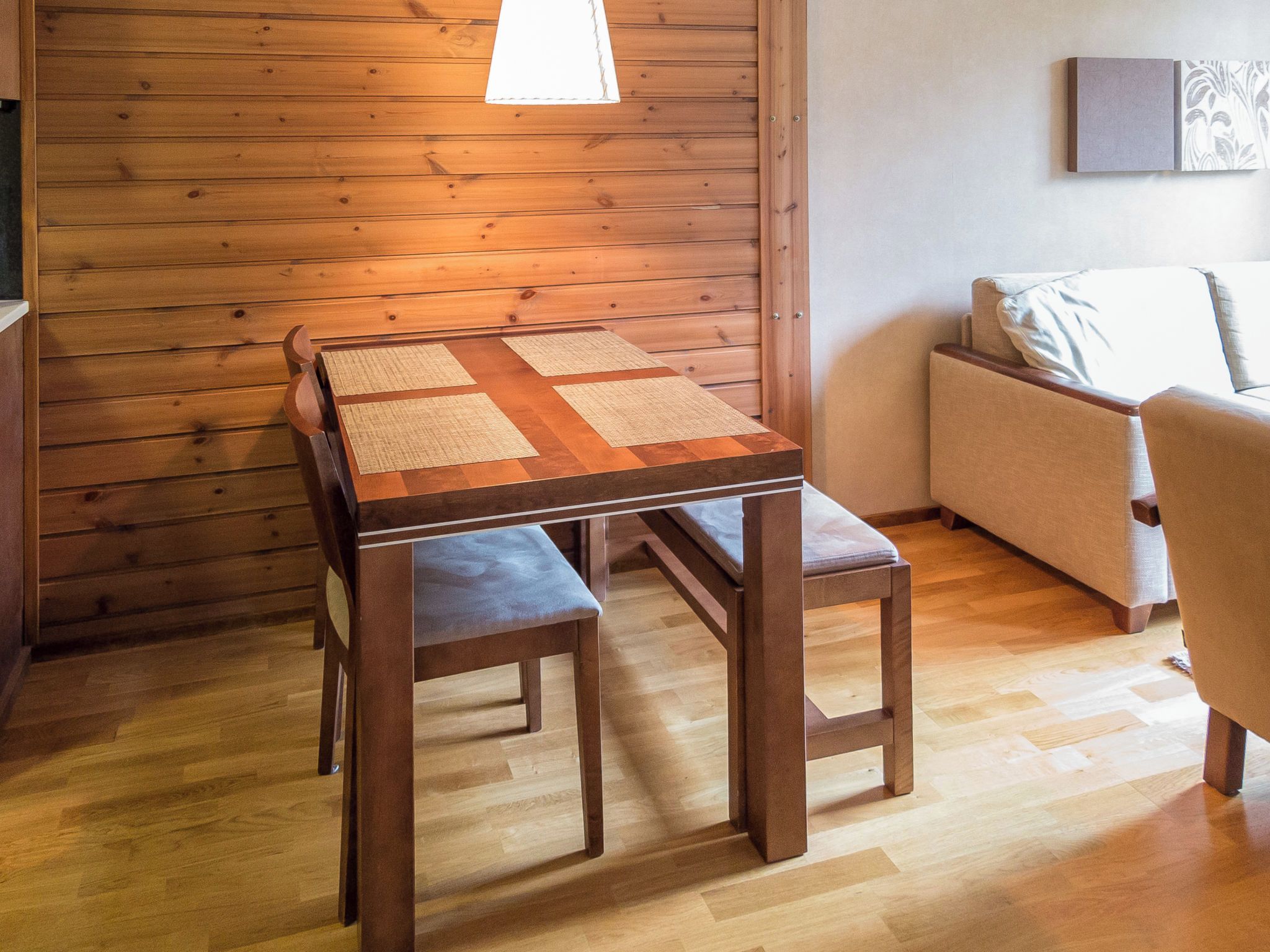 A wooden table with a woven placemat, two matching chairs, and a sofa in a room with wooden panel walls and a light fixture.