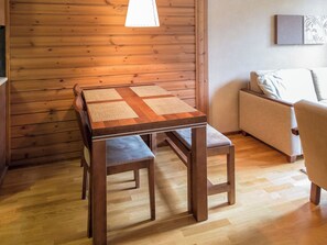 A wooden table with a woven placemat, two matching chairs, and a sofa in a room with wooden panel walls and a light fixture.