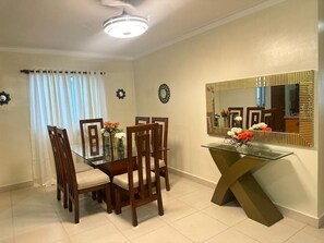 Uma sala de jantar com mesa de vidro, cadeiras de madeira, um espelho e um vaso com flores.