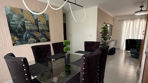 A dining area with a glass table, black upholstered chairs, a potted plant, and a large abstract painting on the wall. - Residencial Palma Real (Puñal)