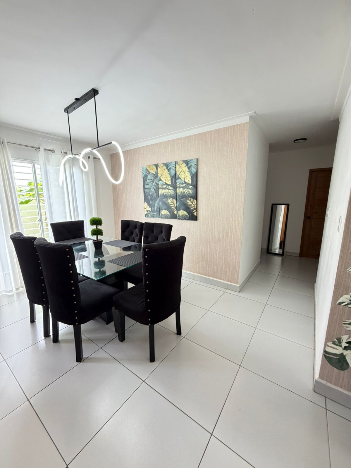 A dining area with a glass table, black chairs, a potted plant, and a wall-mounted painting.