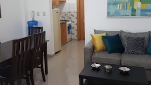Ein Wohnzimmer mit grauem Sofa, einem Couchtisch und einem Essbereich mit Holztisch und Stühlen. Im Hintergrund ist eine Küche mit Holzschränken und einer gefliesten Spritzwand zu sehen.