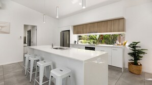 A modern kitchen with a large island, white bar stools, a stainless steel refrigerator, and a window with a view of greenery. - 32c continuously heated pool 300m to beach Seaside@PalmCove (palm cove)