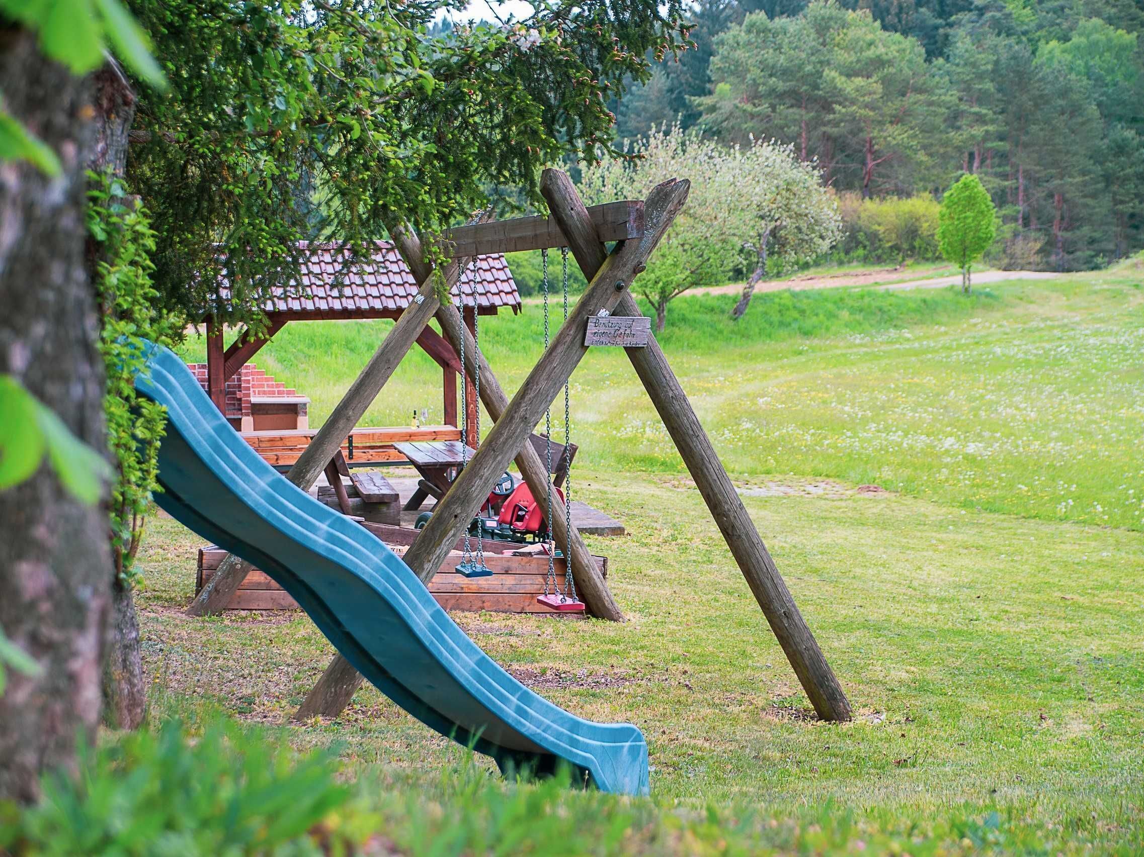 Une aire de jeux avec un toboggan et des balançoires, des tables de pique-nique et un petit bâtiment avec un toit rouge.