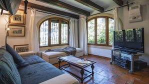 Una sala de estar acogedora con un sofá, una mesa de centro y un mueble de televisión con un televisor de pantalla plana. Hay ventanas amplias con cortinas, un radiador y cuadros enmarcados en la pared.