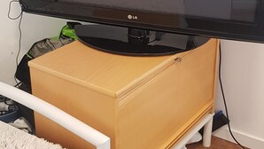 Un televisor sobre un mueble bajo, una cama con cabecera blanca y un pequeño armario de madera.