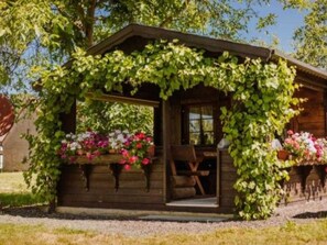 A wooden cabin with a porch covered in green vines, a wooden bench, and flower planters. - Ferienwohnung Erlwein (Eggolsheim)