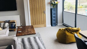 A modern living room with a sofa, a coffee table, a yellow bean bag, a TV mounted on the wall, and a large window offering a view of the outdoors. - Alnmouth Close to Beach and golf with Private Parking and Pet Friendly (Alnwick)