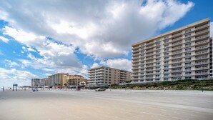 Hotel aan het strand met meerdere verdiepingen, balkons en een zandstrand op de voorgrond.