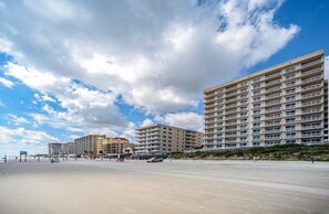 Un hôtel situé en bord de mer, avec plusieurs étages, des balcons et une plage de sable au premier plan.