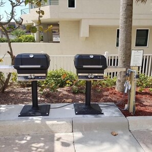 Two outdoor barbecue grills mounted on a concrete base, with a palm tree and a building in the background. - #1005 Sanibel Direct Oceanfront SE Corner Unit 3/3 (Daytona Beach Shores)