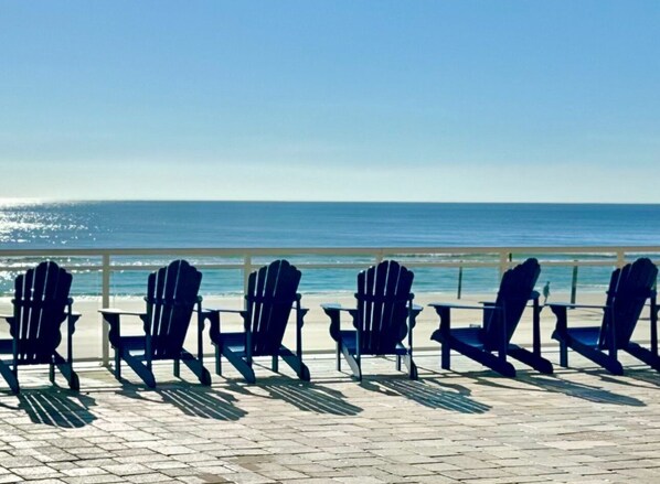 A beachside view with four Adirondack chairs aligned on a paved walkway, overlooking the ocean.
