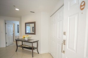 A hallway with a white door, a mirror, a small table with a vase of flowers, and a tiled floor.