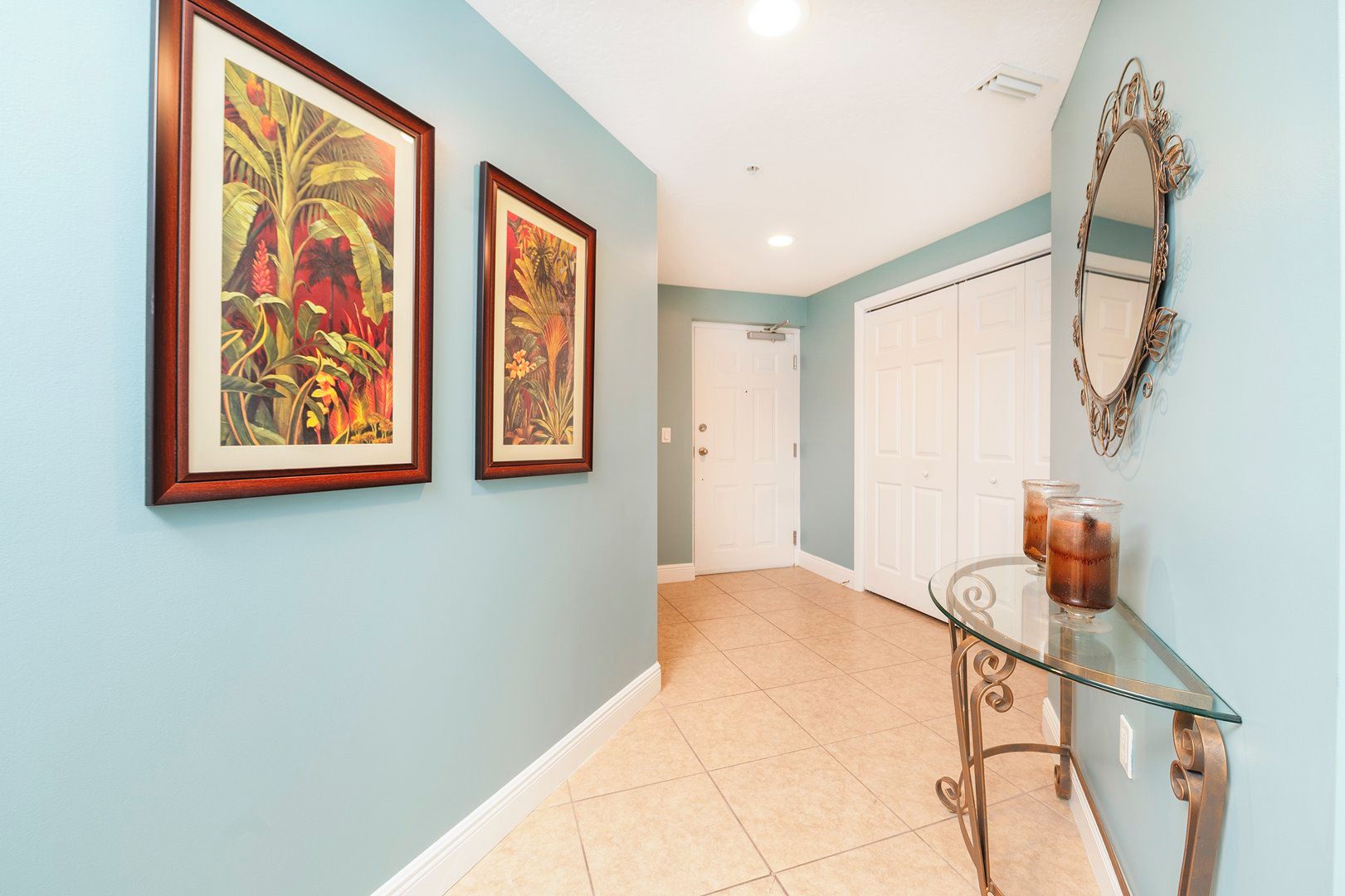 A hallway with light blue walls, two framed tropical paintings, a glass table with drinks, and a decorative mirror.