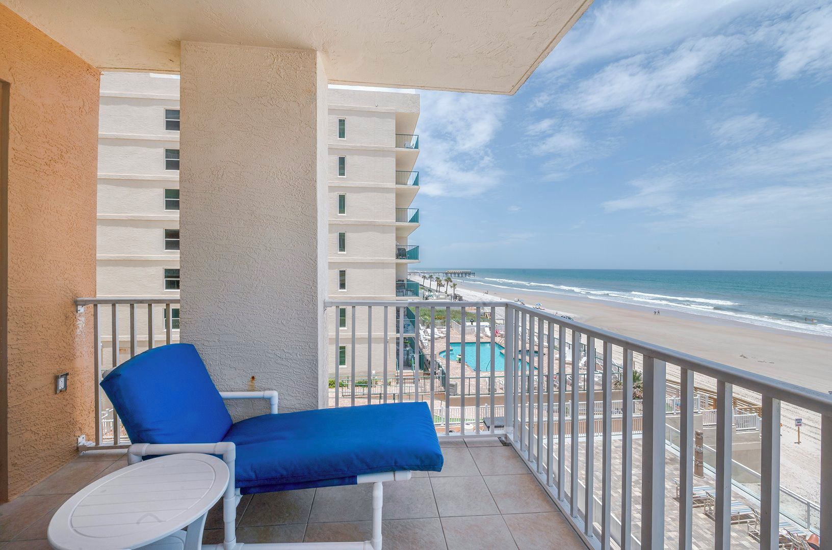 A balcony with a blue chair and a small table, overlooking a beach with a pool and a pier.