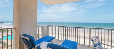 Een balkon met twee loungesteilen en een kleine tafel, met uitzicht op een strand en een zwembad.