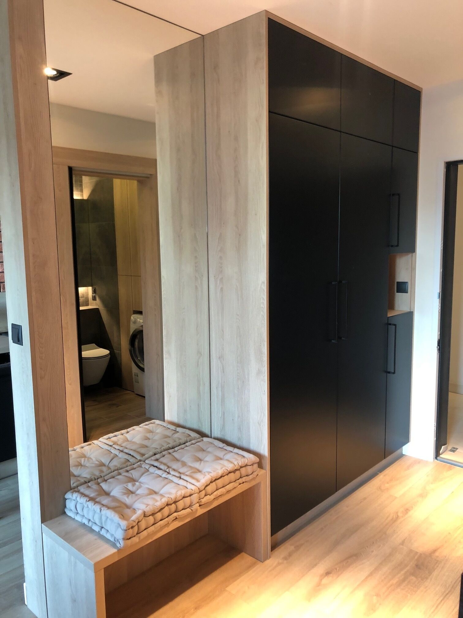 A modern bedroom with a built-in bench, a dark wardrobe, and a visible bathroom with a toilet and washing machine.