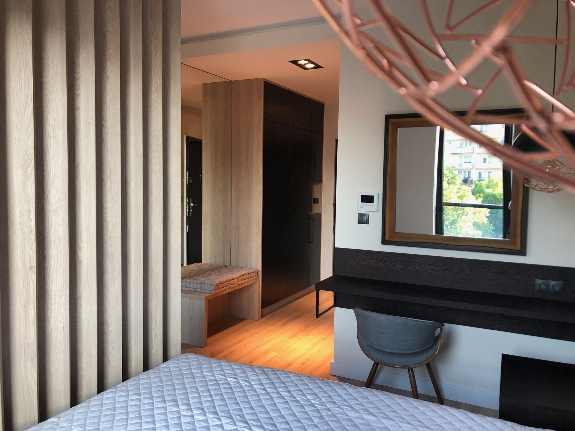 A modern bedroom with a bed, a desk, and a chair. There is a window with a view of buildings and trees.