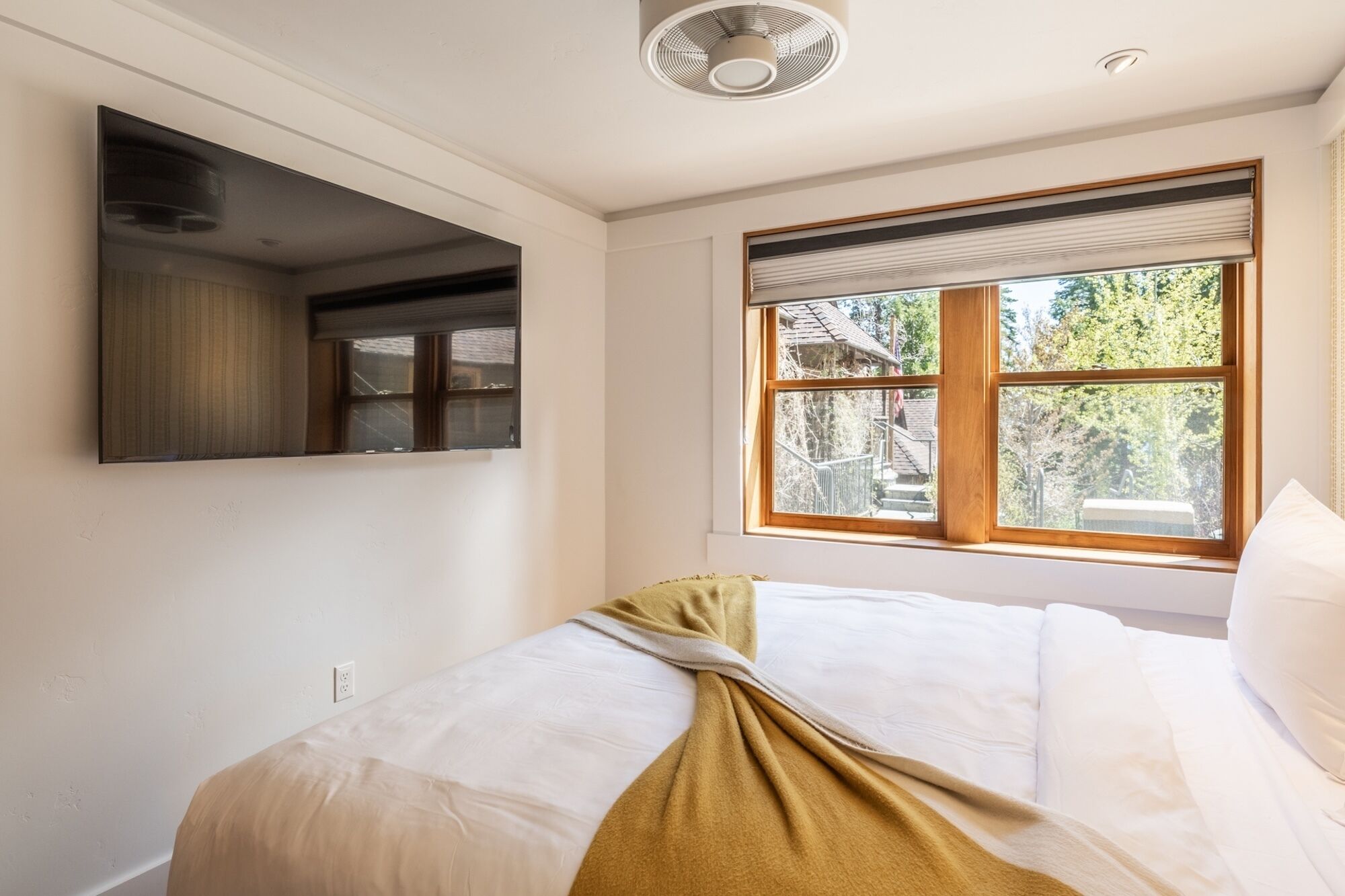 Une chambre à coucher avec un lit, une fenêtre avec des stores, un ventilateur de plafond et un téléviseur fixé au mur.
