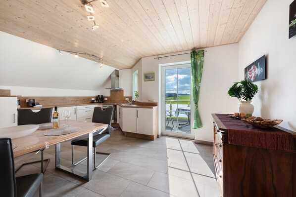 A modern kitchen with a wooden ceiling, a dining table with chairs, a kitchen island, and a view through a glass door.
