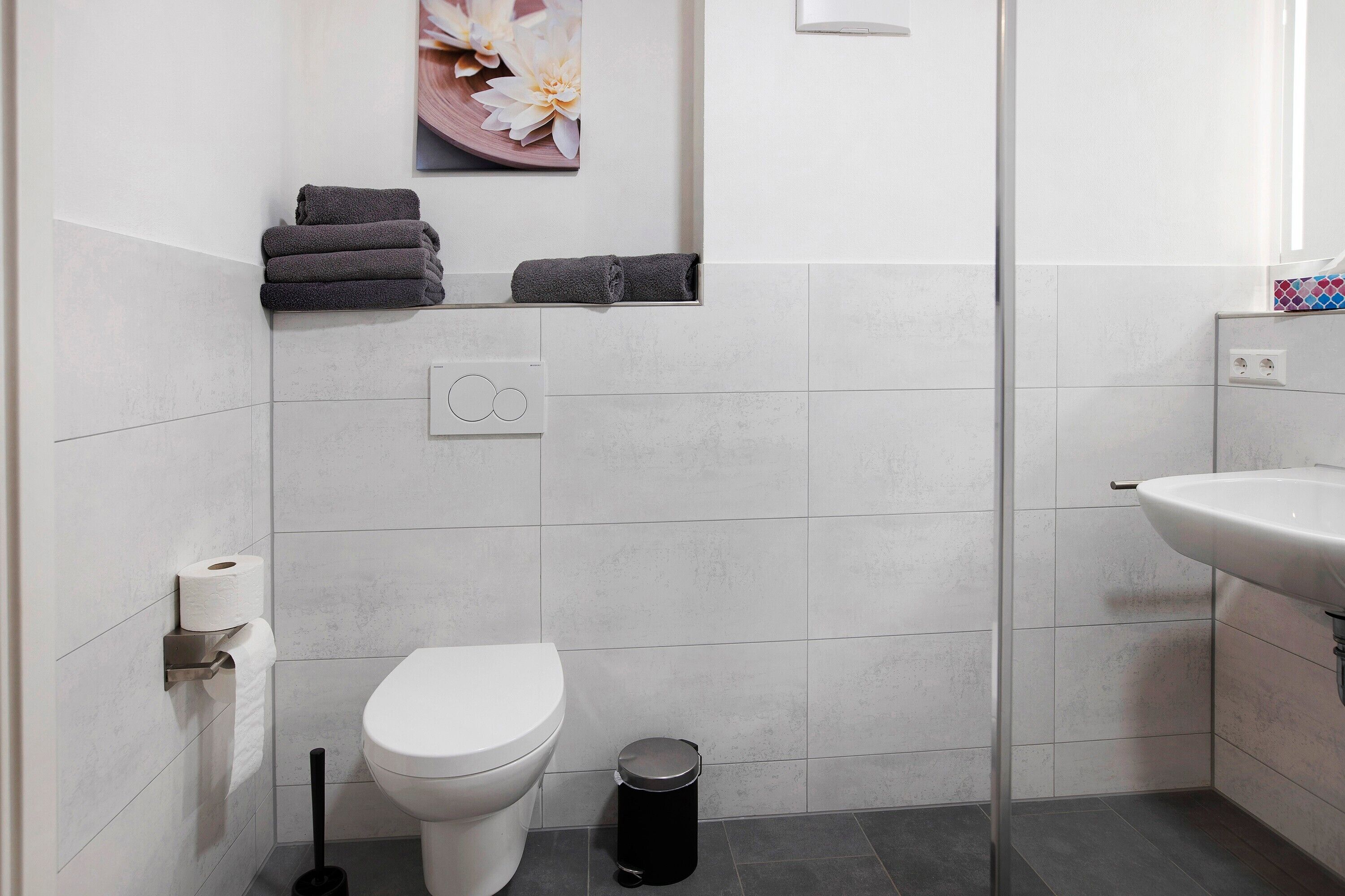 A bathroom with a toilet, a trash bin, a towel rack with folded towels, a toilet paper holder, and a wall-mounted light fixture.