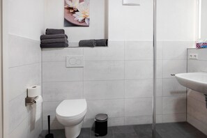Een badkamer met toilet, een prullenbak, een handdoekrek met opgevouwen handdoeken, een toiletrolhouder en een wandlamp.