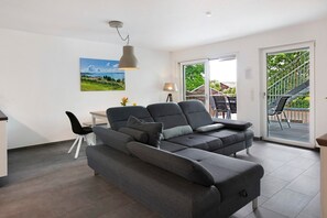 Una sala moderna con un sofá seccional gris, una mesa de comedor y una silla. Hay un ventanal con vistas a zonas verdes y una escalera que lleva a un espacio exterior.