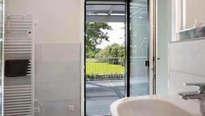 Une salle de bain avec un lavabo blanc, un porte-serviettes et une porte en verre donnant sur un espace extérieur agrémenté d’une clôture en métal et de verdure.