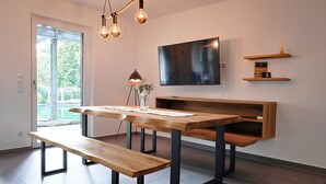 A modern living room with a wooden table, benches, a TV, and a fireplace. - Holiday Apartment "Bodensee" with Terrace, Shared Garden & Swimming Pond (Meßkirch)